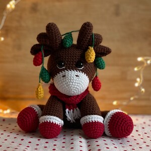 Crochet Reindeer, dark brown in color with white muzzle, he is wearing red and white Christmas shoes on all 4 legs, he has red and white scarfs around his neck and colorful Christmassy lights hanging on his antlers. He is 13sc tall then sitting.