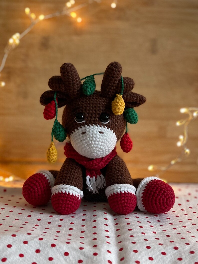 Crochet Reindeer, dark brown in color with white muzzle, he is wearing red and white Christmas shoes on all 4 legs, he has red and white scarfs around his neck and colorful Christmassy lights hanging on his antlers. He is 13sc tall then sitting.