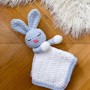 Puede incluir: Una manta de crochet azul y blanca con forma de conejo. El conejo est&aacute; acostado con los ojos cerrados y tiene la nariz rosa. La manta est&aacute; hecha de un hilo suave y esponjoso.