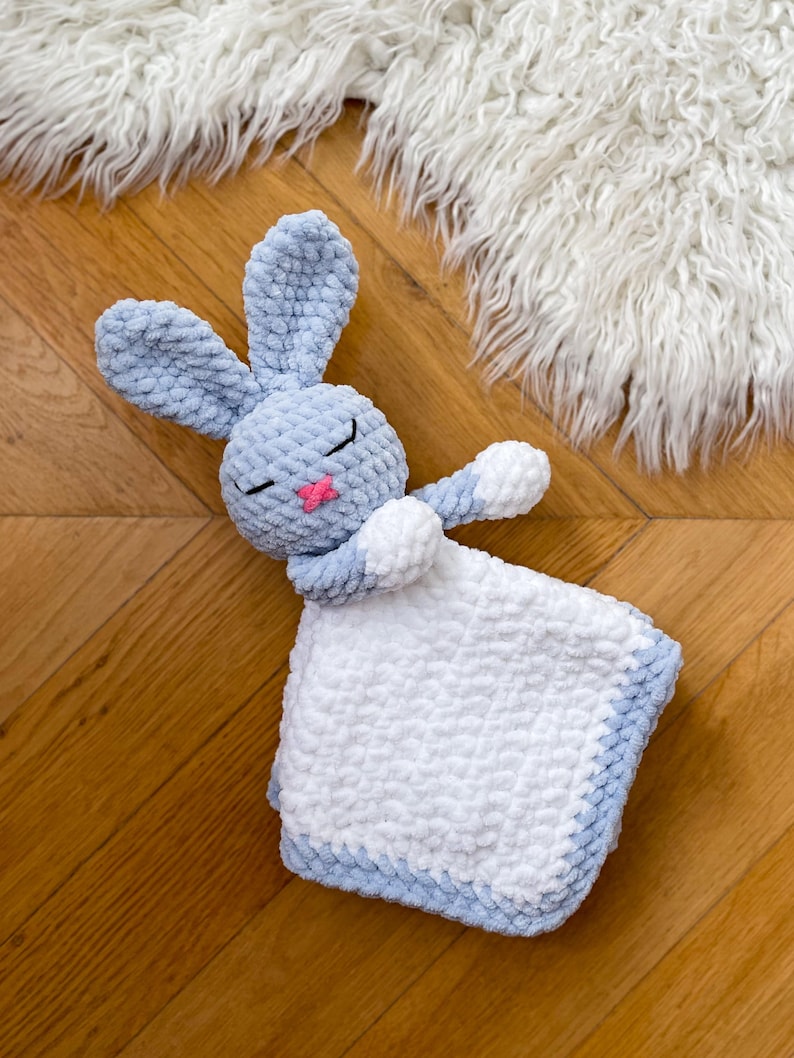 Puede incluir: Una manta de crochet azul y blanca con forma de conejo. El conejo est&aacute; acostado con los ojos cerrados y tiene la nariz rosa. La manta est&aacute; hecha de un hilo suave y esponjoso.