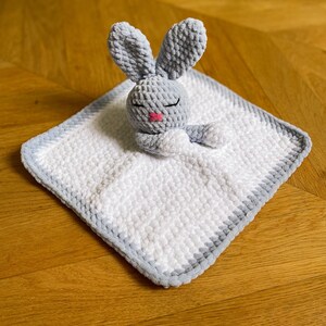 Puede incluir: Un conejito de crochet gris con la nariz rosa y los ojos cerrados est&aacute; acostado sobre una manta de beb&eacute; de crochet blanca y gris.