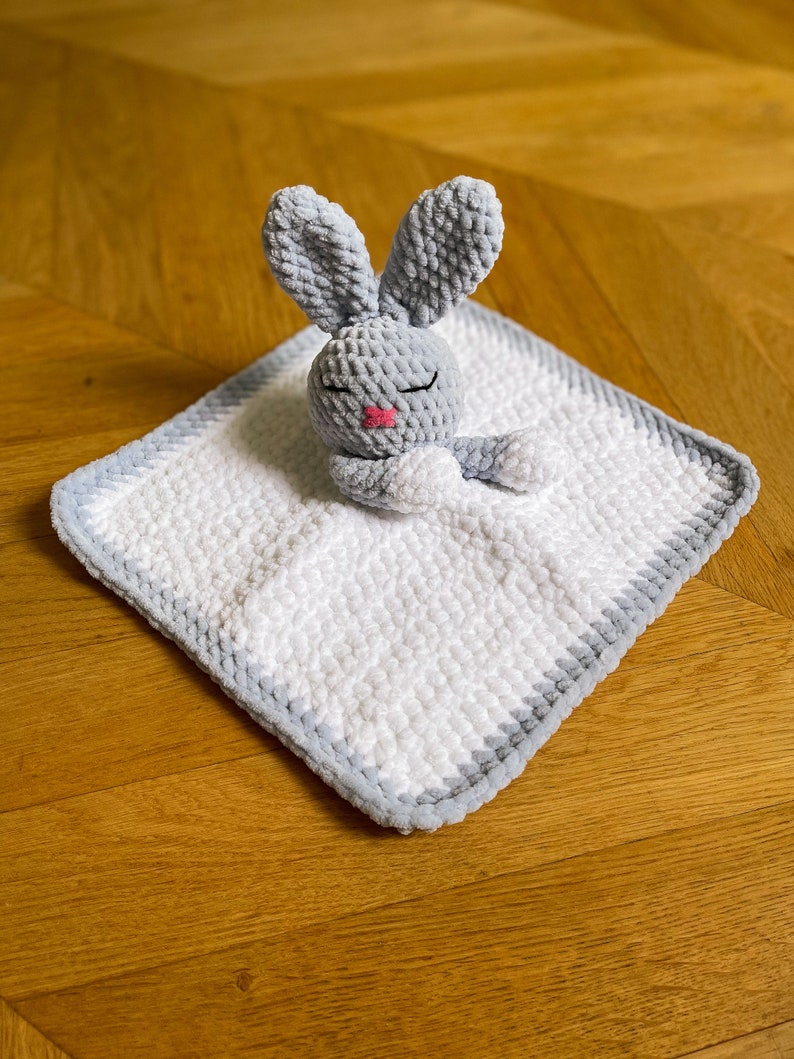 Puede incluir: Un conejito de crochet gris con la nariz rosa y los ojos cerrados est&aacute; acostado sobre una manta de beb&eacute; de crochet blanca y gris.