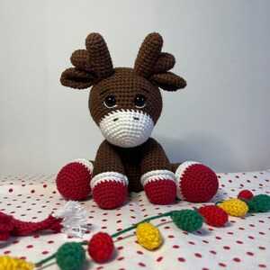 Crochet Reindeer, dark brown in color with white muzzle, he is wearing red and white Christmas shoes on all 4 legs, he has red and white scarfs around his neck and colorful Christmassy lights hanging on his antlers. He is 13sc tall then sitting.