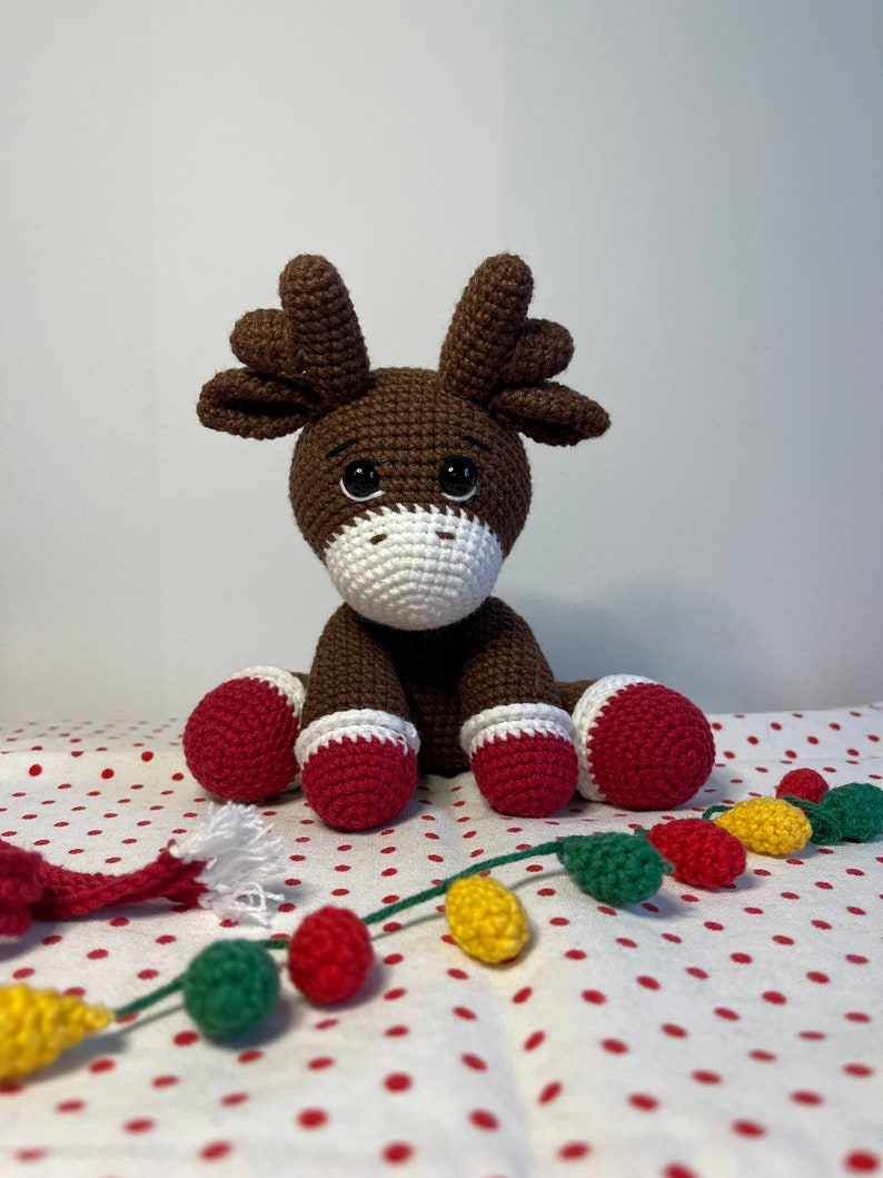 Crochet Reindeer, dark brown in color with white muzzle, he is wearing red and white Christmas shoes on all 4 legs, he has red and white scarfs around his neck and colorful Christmassy lights hanging on his antlers. He is 13sc tall then sitting.