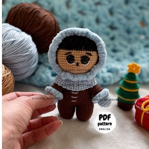 Puede incluir: Mu&ntilde;eca amigurumi de crochet con un atuendo de invierno azul con capucha y pantalones marrones. La mu&ntilde;eca tiene cabello negro y ojos marrones. La mu&ntilde;eca est&aacute; de pie sobre una manta azul y blanca con un peque&ntilde;o &aacute;rbol de Navidad y una caja de regalo roja en el fondo. El texto "PDF pattern ENG/SK" est&aacute; en un c&iacute;rculo blanco.
