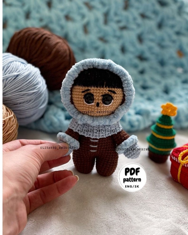 Puede incluir: Mu&ntilde;eca amigurumi de crochet con un atuendo de invierno azul con capucha y pantalones marrones. La mu&ntilde;eca tiene cabello negro y ojos marrones. La mu&ntilde;eca est&aacute; de pie sobre una manta azul y blanca con un peque&ntilde;o &aacute;rbol de Navidad y una caja de regalo roja en el fondo. El texto "PDF pattern ENG/SK" est&aacute; en un c&iacute;rculo blanco.