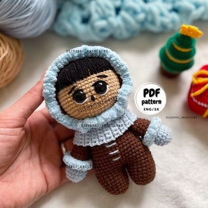 Puede incluir: Mu&ntilde;eca amigurumi de crochet con un abrigo con capucha azul y pantalones marrones. La mu&ntilde;eca tiene el pelo negro y los ojos marrones. La mu&ntilde;eca sostiene una manopla de crochet azul. La imagen incluye el texto "PDF pattern ENG/SK".