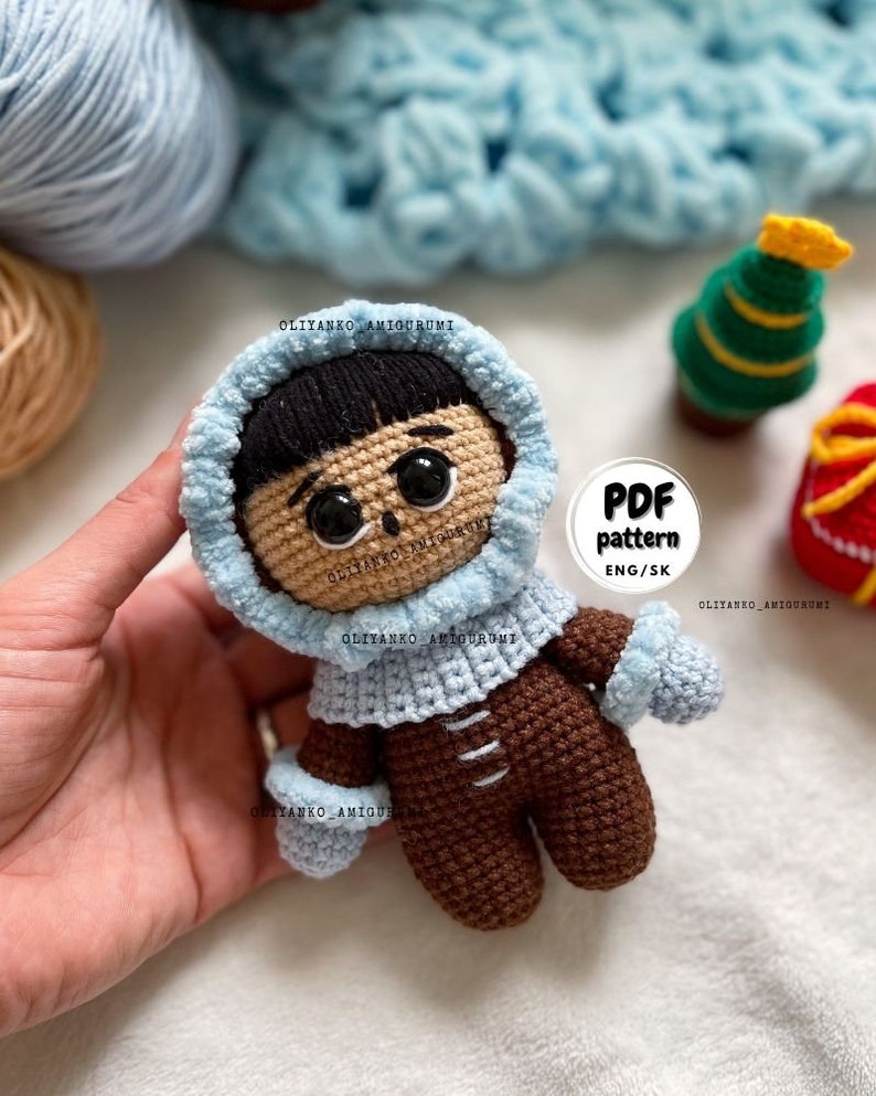 Puede incluir: Mu&ntilde;eca amigurumi de crochet con un abrigo con capucha azul y pantalones marrones. La mu&ntilde;eca tiene el pelo negro y los ojos marrones. La mu&ntilde;eca sostiene una manopla de crochet azul. La imagen incluye el texto "PDF pattern ENG/SK".