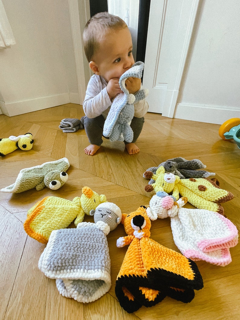 Puede incluir: Una colecci&oacute;n de mantas de beb&eacute; de crochet con caras de animales. Las mantas son de varios colores, incluyendo amarillo, naranja, gris y blanco. Las mantas est&aacute;n dispuestas en un suelo de madera.