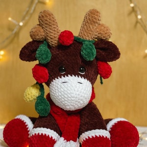 May include: A brown crocheted reindeer toy with red and green holly accents, wearing a red scarf. The reindeer is sitting on a white fluffy surface with string lights in the background.