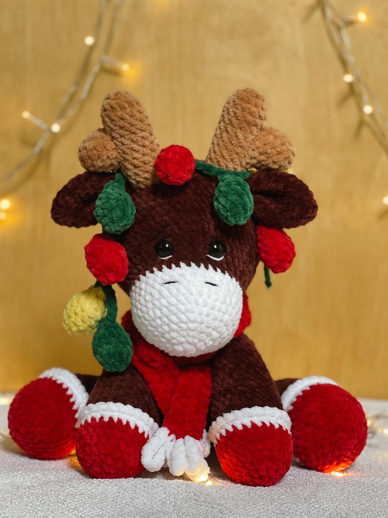 May include: A brown crocheted reindeer toy with red and green holly accents, wearing a red scarf. The reindeer is sitting on a white fluffy surface with string lights in the background.