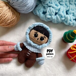 Puede incluir: Mu&ntilde;eca amigurumi de crochet con una chaqueta con capucha azul, pantalones marrones y guantes azules. La mu&ntilde;eca tiene cabello casta&ntilde;o y ojos grandes. La mu&ntilde;eca est&aacute; sentada sobre una superficie blanca con una manta de crochet azul, una bola de hilo marr&oacute;n y una bola de hilo azul en el fondo. La imagen tambi&eacute;n incluye un peque&ntilde;o &aacute;rbol de Navidad de crochet y una caja de regalo roja y dorada de crochet. Un c&iacute;rculo blanco con texto negro dice "PDF pattern ENG/SK".