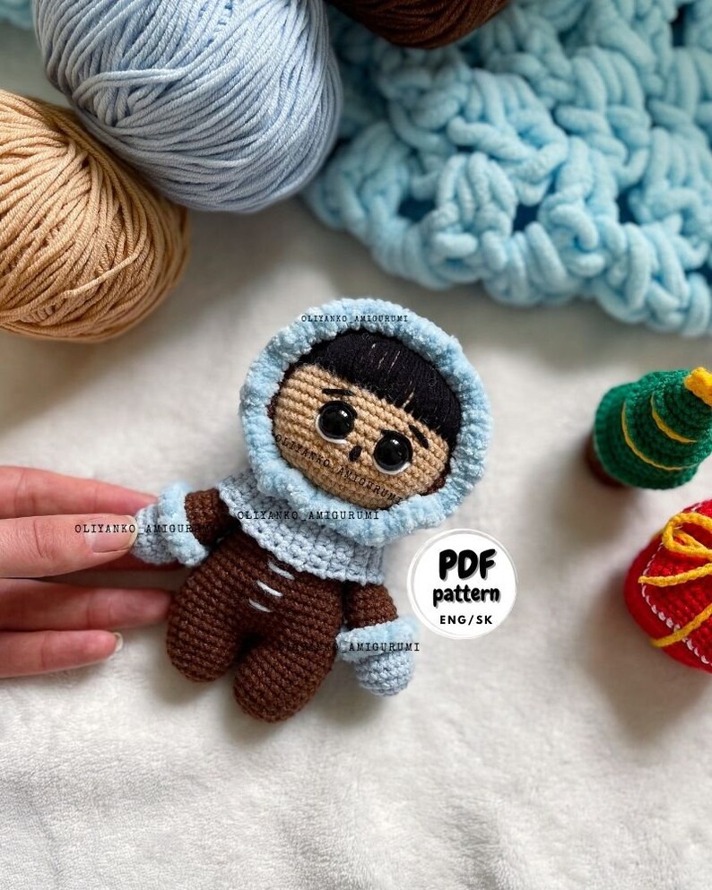 Puede incluir: Mu&ntilde;eca amigurumi de crochet con una chaqueta con capucha azul, pantalones marrones y guantes azules. La mu&ntilde;eca tiene cabello casta&ntilde;o y ojos grandes. La mu&ntilde;eca est&aacute; sentada sobre una superficie blanca con una manta de crochet azul, una bola de hilo marr&oacute;n y una bola de hilo azul en el fondo. La imagen tambi&eacute;n incluye un peque&ntilde;o &aacute;rbol de Navidad de crochet y una caja de regalo roja y dorada de crochet. Un c&iacute;rculo blanco con texto negro dice "PDF pattern ENG/SK".