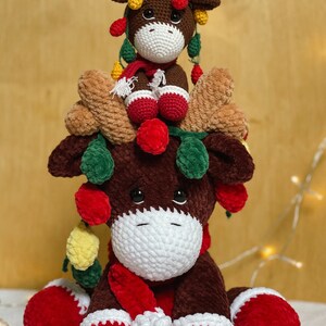 May include: Two crocheted reindeer ornaments, one sitting on top of the other. The larger reindeer is brown with a red scarf and white trim. The smaller reindeer is brown with a red scarf and white trim. Both reindeer have green and yellow lights around their antlers.