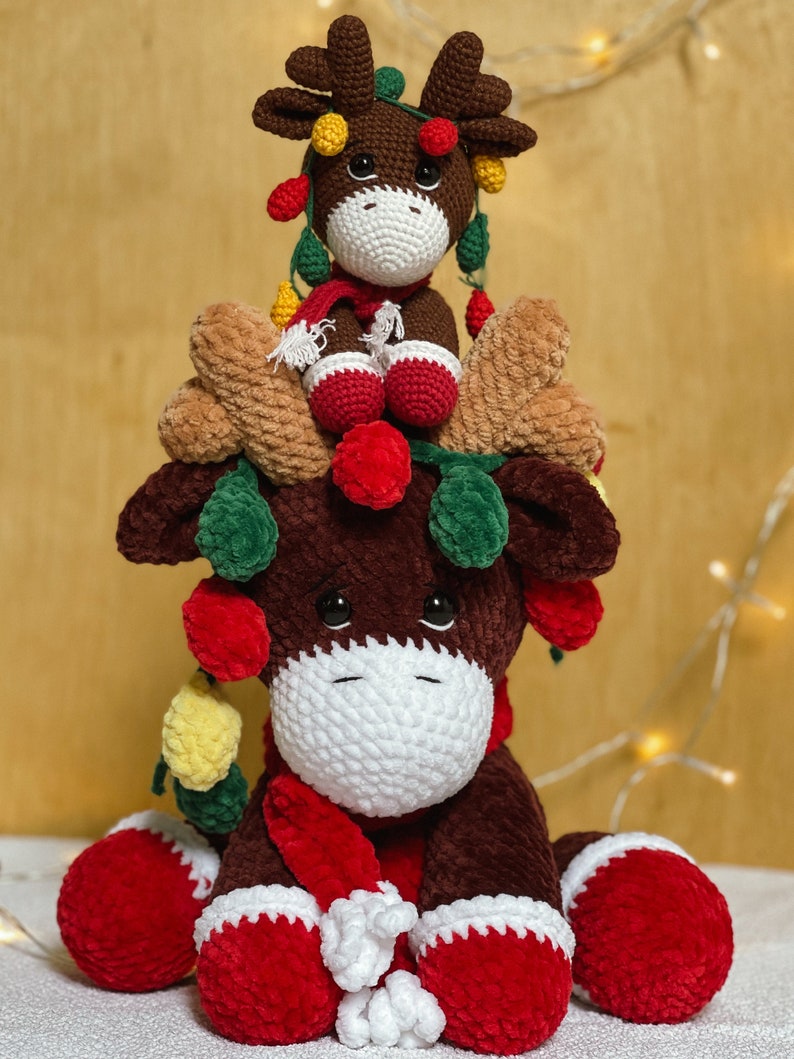 May include: Two crocheted reindeer ornaments, one sitting on top of the other. The larger reindeer is brown with a red scarf and white trim. The smaller reindeer is brown with a red scarf and white trim. Both reindeer have green and yellow lights around their antlers.