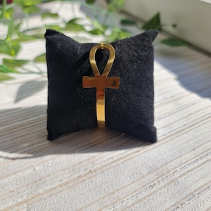May include: A gold-colored Ankh bracelet displayed on a black velvet pillow. The Ankh symbol, a cross with a loop at the top, is the focal point. The bracelet is made of a shiny, reflective metal. The background is a light-colored surface with green foliage.
