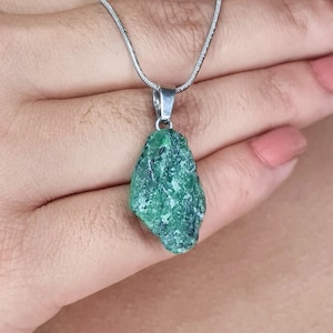 May include: A silver necklace featuring a raw, emerald-green gemstone pendant. The pendant has a rough, natural texture and is suspended from a delicate silver chain. The gemstone is a vibrant shade of green, with darker inclusions.