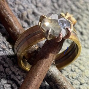 Può includere: Un anello multicolore con una grande gemma trasparente sfaccettata. L'anello presenta una fascia dorata con accenti in rame e argento. La gemma è tenuta in posizione da griffe d'oro. L'anello è esposto su un rametto marrone.