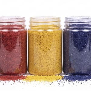 May include: A row of seven clear plastic jars filled with colorful, granular material. The jars contain blue, green, red, yellow, dark blue, purple, and teal substances. A small amount of each color is scattered below the jars on a white surface.