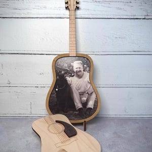 Puede incluir: Marco de fotos de madera con forma de guitarra con una imagen en blanco y negro de un hombre y un perro. Un marco de fotos más pequeño con forma de guitarra está delante. Se ve el clavijero y el mástil de la guitarra. El marco está sobre un soporte de madera.