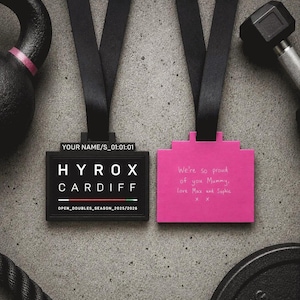 Pode incluir: Duas medalhas com fitas pretas são exibidas contra um fundo cinza. Uma é preta com o texto "HYROX CARDIFF", a outra é rosa com texto manuscrito. Equipamentos de fitness, incluindo um kettlebell e um haltere, estão ao fundo.