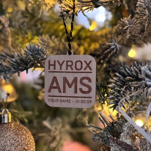 May include: A wooden Christmas bauble with the text "HYROX AMS" and "YOUR NAME - 01:00:59" hanging on a Christmas tree.