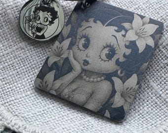 Puede incluir: Un llavero de metal con un colgante cuadrado y un dije circular más pequeño, ambos con una ilustración en escala de grises de Betty Boop rodeada de flores de lirio. El llavero incluye una argolla plateada estándar.