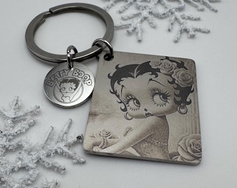 Puede incluir: Un llavero de metal que presenta un colgante rectangular con una ilustración en tono sepia de Betty Boop y un dije circular más pequeño grabado con el texto Betty Boop. El llavero incluye una anilla estándar para sujeción.