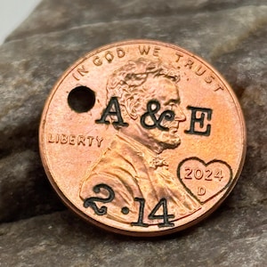 May include: A copper penny with the image of Abraham Lincoln, featuring the text "IN GOD WE TRUST" and "LIBERTY." The penny is stamped with the initials "A & E," the date "2024," and the numbers "2.14" with a heart symbol.