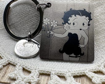 Puede incluir: Un llavero de metal con un colgante cuadrado que muestra una ilustración en blanco y negro de Betty Boop sosteniendo flores, y un dije circular más pequeño grabado con un dibujo lineal de Betty Boop y un perro dentro de un corazón.