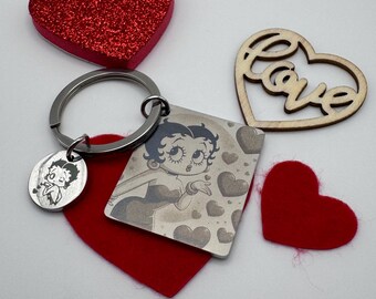 Puede incluir: Un llavero de metal con un dije cuadrado que muestra una imagen impresa de Betty Boop lanzando un beso rodeada de corazones, y un dije circular más pequeño con una ilustración en blanco y negro de Betty Boop, junto a corazones rojos y de madera.