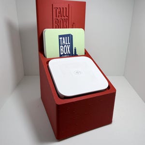 May include: A red 3D-printed stand featuring the embossed text TALL BOX and a lighthouse icon. It holds a square white payment terminal and a light green card with the text TALL BOX GAMES and a lighthouse icon printed in dark blue ink.
