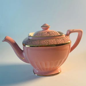 May include: A pastel pink ceramic teapot featuring a gold-trimmed lid and upper body with a textured, honeycomb-like gold pattern. The teapot has a curved spout, a matching handle, and a fluted base design with gold accents along the edges.