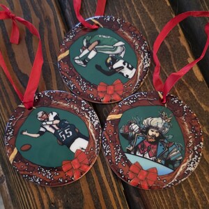 May include: Three round ceramic ornaments with a brown and white pretzel design. Each ornament features a different image: a football player, a football player making a catch, and a man in a blue and white costume with a red bow tie. Each ornament has a red ribbon attached.