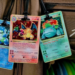 May include: Three vintage Pokemon trading cards featuring Charizard, Blastoise, and Venusaur. The cards are holographic and have colorful illustrations of the Pokemon. The cards are attached to a brown bag with a black strap.