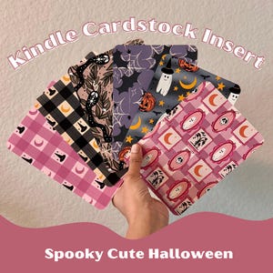 Könnte beinhalten: Eine Hand hält eine Auswahl an Kindle-Cardstock-Einsätzen mit Halloween-Mustern. Designs umfassen Fledermäuse, Geister, Kürbisse, Monde und Totenköpfe in Rosa, Schwarz und Lila. Der Text lautet "Kindle Cardstock Insert" und "Spooky Cute Halloween".