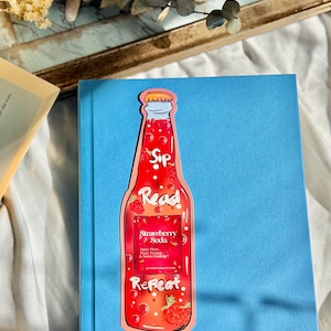 Pu&ograve; includere: Un segnalibro rosso e rosa a forma di bottiglia di soda alla fragola. La bottiglia ha un'etichetta bianca che dice "Sip Read Repeat" e "Strawberry Soda" con il testo "Juicy Plots, Happy Tears & Sweet Ending!" sotto.