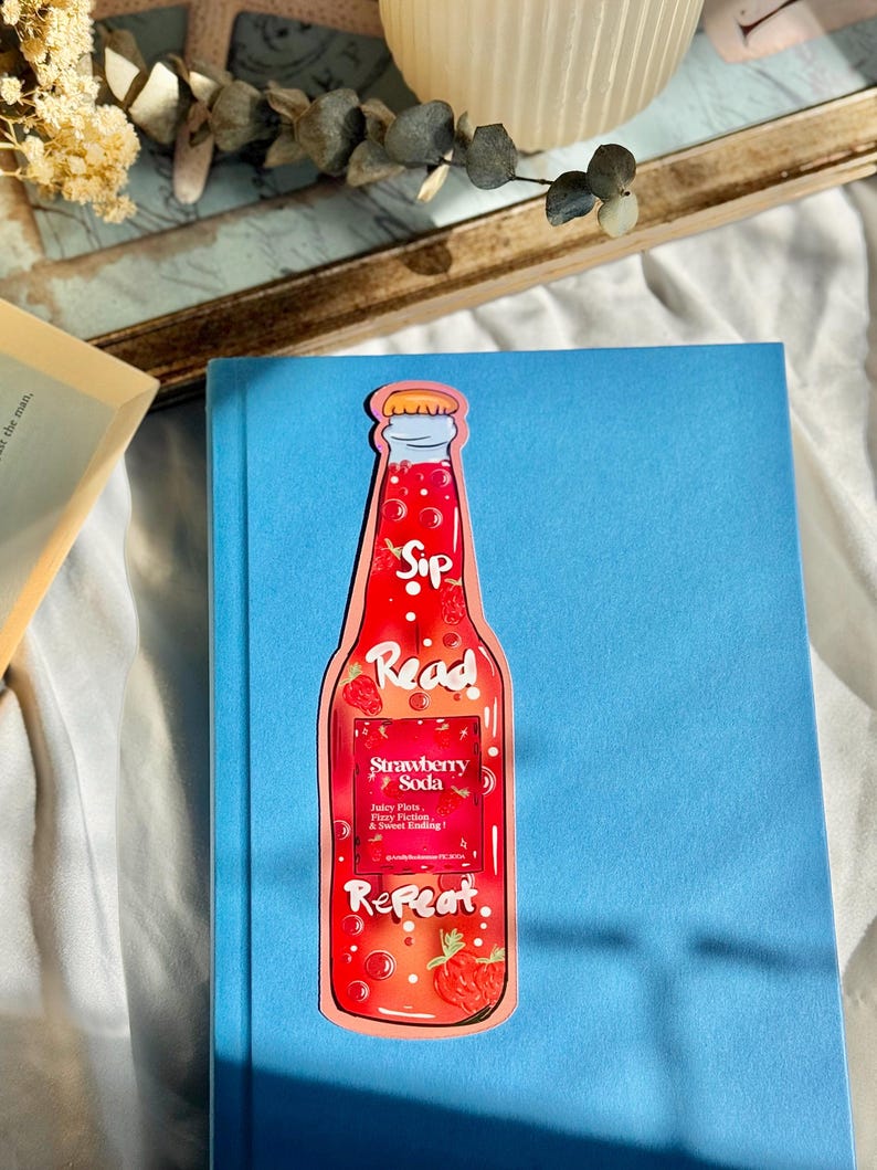 Pu&ograve; includere: Un segnalibro rosso e rosa a forma di bottiglia di soda alla fragola. La bottiglia ha un'etichetta bianca che dice "Sip Read Repeat" e "Strawberry Soda" con il testo "Juicy Plots, Happy Tears & Sweet Ending!" sotto.
