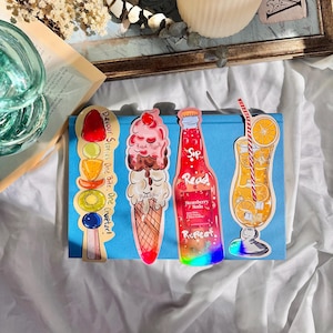 Pu&ograve; includere: Tre segnalibri colorati che raffigurano una bottiglia di soda alla fragola, una pallina di gelato in un cono di cialda e un bicchiere di succo d'arancia. I segnalibri sono stampati con il testo "Sip Read Repeat" e "Devour Stories One Bite, One Chapter!"