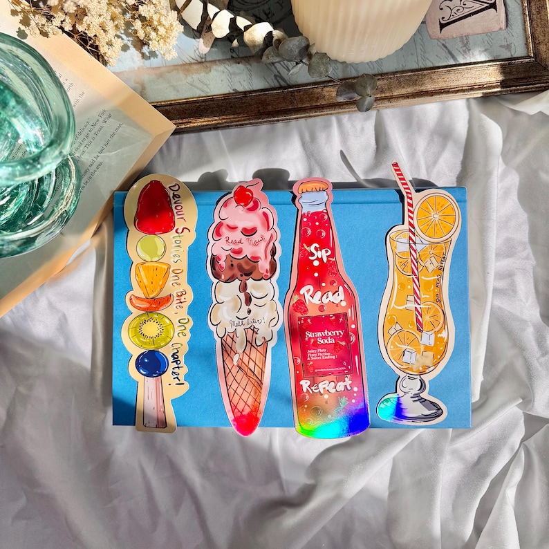 Pu&ograve; includere: Tre segnalibri colorati che raffigurano una bottiglia di soda alla fragola, una pallina di gelato in un cono di cialda e un bicchiere di succo d'arancia. I segnalibri sono stampati con il testo "Sip Read Repeat" e "Devour Stories One Bite, One Chapter!"