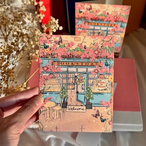 Könnte beinhalten: Eine Grußkarte mit einer Buchladen-Illustration, rosa Blumen, Schmetterlingen und dem Wort "Welcome". Die Karte hat ein rosa und blaues Farbschema, mit dem Wort "Bookstore" oben. Weitere Bücher sind im Hintergrund zu sehen.