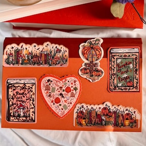 May include: A selection of autumn-themed stickers with pumpkins, books, and mushrooms. The stickers are in various shapes, including a heart and rectangular shapes. The colour palette includes orange, brown, and green.