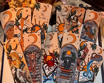 May include: A collection of illustrated zodiac sign bookmarks. Each bookmark features a unique design with astrological symbols, crescent moons, and botanical elements. The color palette includes orange, beige, and blue tones. The bookmarks are arranged in a fan shape.