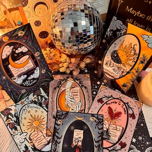 May include: An assortment of tarot cards with celestial and botanical illustrations, including a sun, moon, and open book. The cards are arranged around a disco ball and dried flowers, creating a mystical aesthetic. The text on the cards includes "Morning Reader", "Fated Mate", and "Enemies to Lovers".