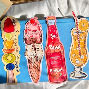 Pu&ograve; includere: Tre segnalibri colorati con uno spiedino di frutta, un cono gelato e una bottiglia di soda alla fragola. I segnalibri sono progettati con un tema estivo e includono il testo "Devour Stories One Bite, One Chapter!" "Read Now" "Melt later!" "Sip Read" "Strawberry Soda" "Juicy Flavors, Fuzzy Feelings & Sweet Reading" "Repeat."
