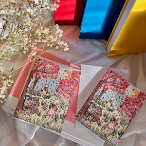 May include: Two greeting cards with a pink and white floral design featuring two bunnies holding baskets of flowers and a picnic basket. The cards are set on a white surface with a pink and white floral design.