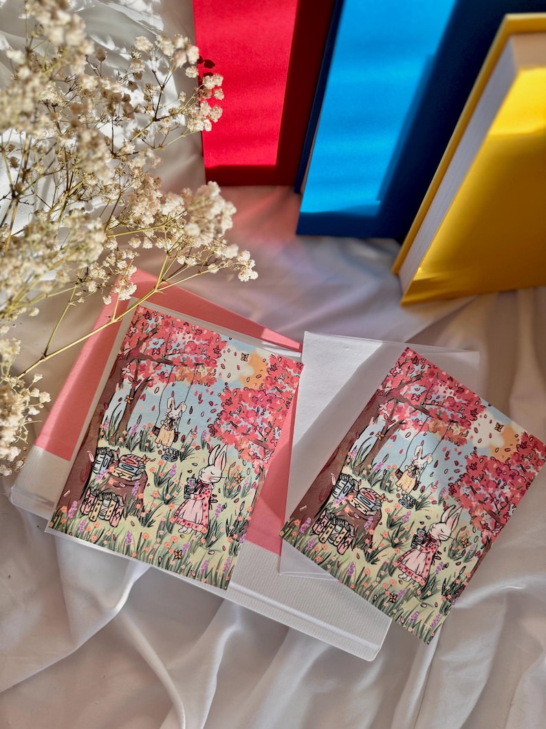 May include: Two greeting cards with a pink and white floral design featuring two bunnies holding baskets of flowers and a picnic basket. The cards are set on a white surface with a pink and white floral design.