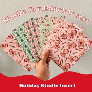 May include: A hand holds a fanned array of holiday-themed Kindle cardstock inserts. The inserts feature festive patterns with Christmas trees, bows, gingerbread men, and teddy bears in red, green, and pink. The text "Kindle Cardstock Insert" and "Holiday Kindle Insert" are visible.