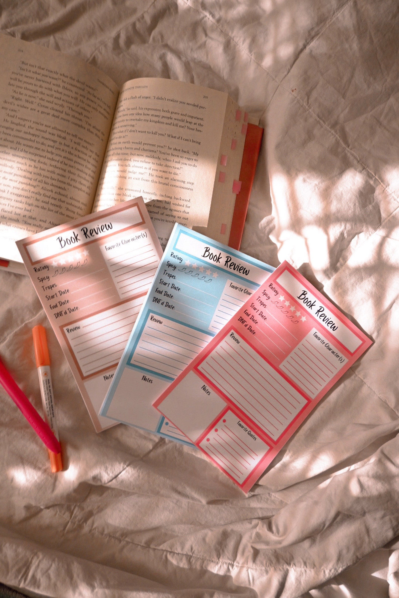 Three book review notepads, tan, blue, and pink. They have sections for notes and reviews and for rankings and favorites. They are on top of a white cloth and an open book.