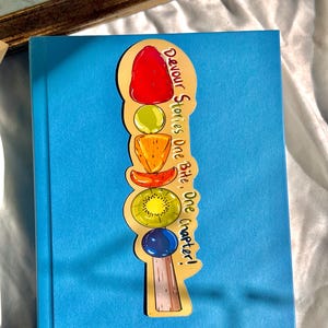Pu&ograve; includere: Un segnalibro dorato con una pila di frutta colorata e il testo "Devour Stories One Bite, One Chapter!"